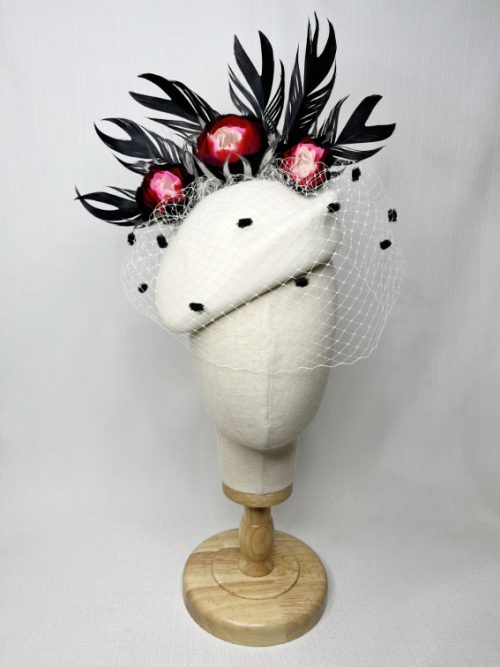 A mannequin wearing a light coloured felt fascinator with feathers and a spotted veil.