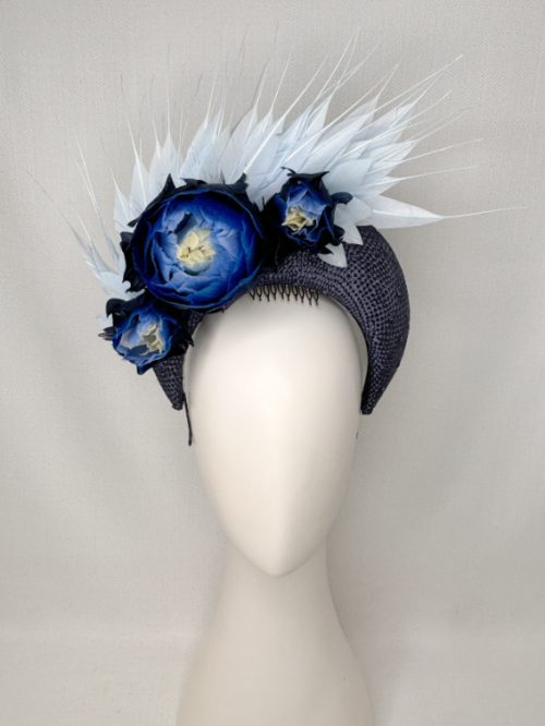 Mannequin head with no face wearing a navy blue fascinator headband with feather flowers and a halo of pale blue diamond cut feathers.