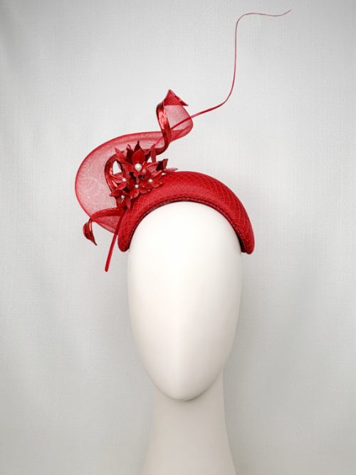 Mannequin head with no face wearing a red fascinator headband with red flowers, a large swirl and curled feather quill.