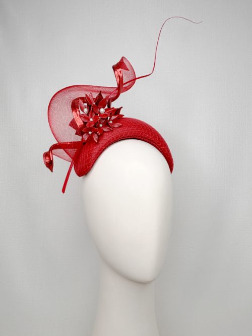 Mannequin head with no face wearing a red fascinator headband with red flowers, a large swirl and curled feather quill.