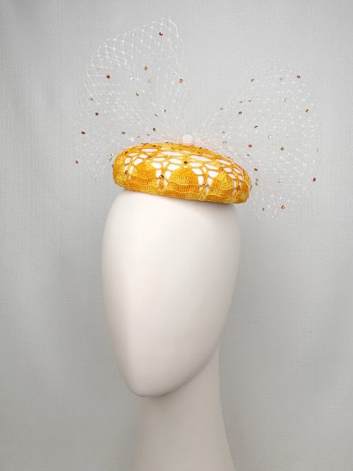 A mannequin head with no face wearing a white and yellow round fascinator with a jewelled bow made from veiling.